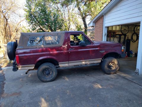 1996 Ford Bronco - A maroon 1996 Ford Bronco with a tan soft top is parked in front of a garage.