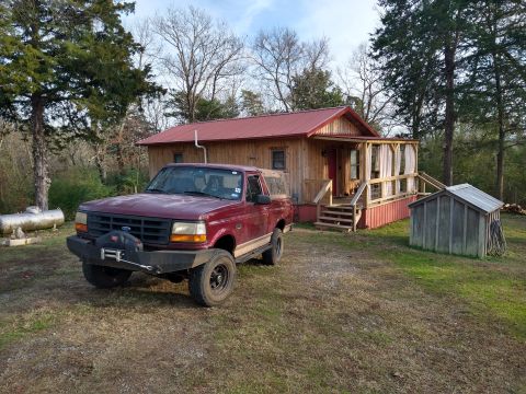 1996 Ford Bronco - A red 1996 Ford Bronco sits parked in front of a rustic wooden house on a grassy lot.