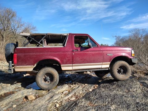 1996 Ford Bronco - A maroon 1996 Ford Bronco with a tan stripe navigates a rocky terrain under a blue sky.
