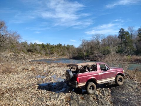 1996 Ford Bronco - A red 1996 Ford Bronco is parked on a rocky area next to a river under a blue sky.