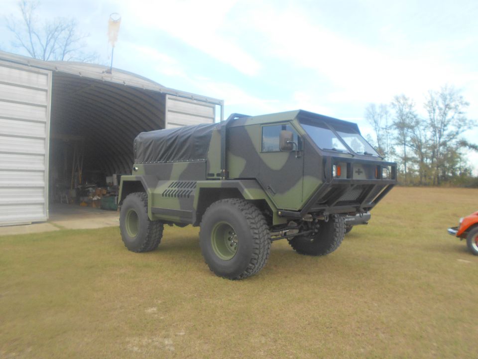 Camouflaged military-style truck with black canvas cover parked on grass in front of a metal building.