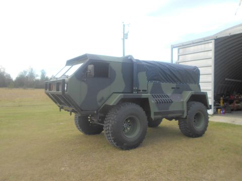 1992 Ford Bronco - A camouflaged, four-wheel-drive truck with a black canvas top is parked on a grassy area next to an open-sided building. The vehicle has la…