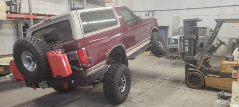 1996 Ford Bronco - A maroon 1996 Ford Bronco is lifted by a forklift, with its rear end elevated high off the ground. Two red jerry cans are mounted to the sp…
