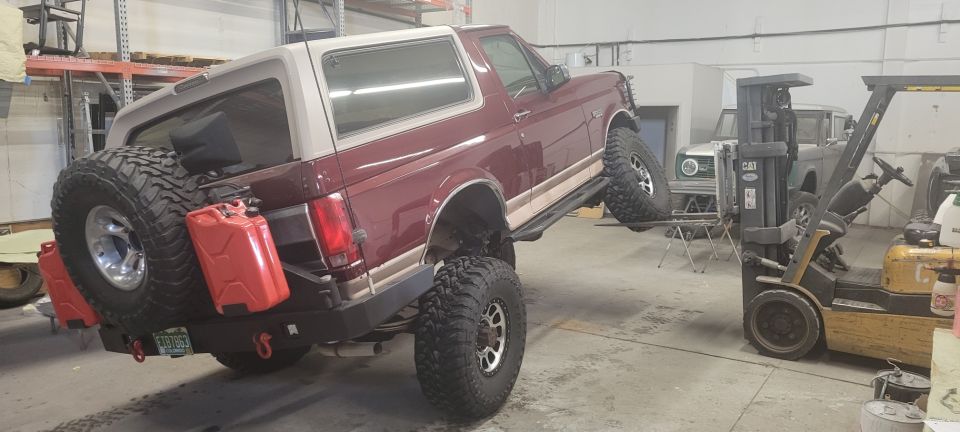 Maroon 1996 Ford Bronco lifted by a forklift, rear end high, with two red jerry cans on the spare tire carrier.