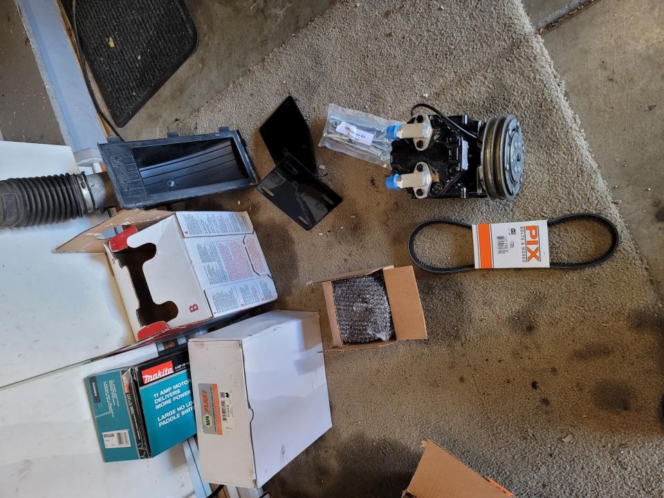 Automotive parts including AC compressor, belt, and air intake hose on carpet.