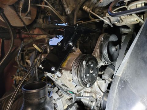 1996 Ford Bronco - A black metal bracket is mounted to the front of a vehicle's engine, securing the air conditioning compressor. The compressor, pulleys, and…