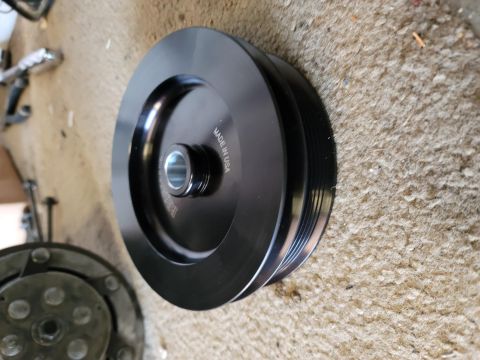 1996 Ford Bronco - A black, multi-groove pulley with "MADE IN USA" engraved on it is shown resting on a carpeted surface. A portion of a metallic component is…
