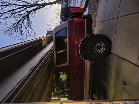 1996 Ford Bronco - A maroon 1996 Ford Bronco with a tan top is parked in a driveway next to a garage. The vehicle has a spare tire mounted on the rear and a r…