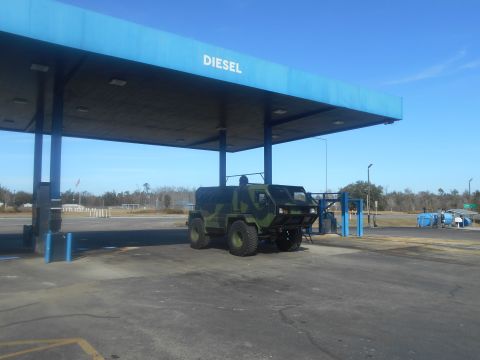 1992 Ford Bronco - A camouflaged, rugged vehicle with large tires is parked under a blue diesel canopy at a gas station. The vehicle has a reinforced front bu…