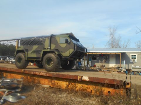 1992 Ford Bronco - A camouflaged, heavy-duty truck with a canvas cover over the bed is parked on a rusty flatbed trailer. A light-colored building with a porc…