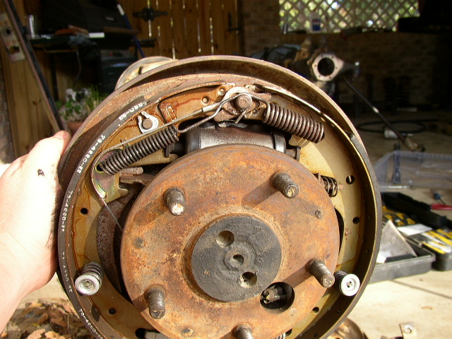 Hand holding a rusty 5-lug drum brake backing plate with shoes and springs in a garage
