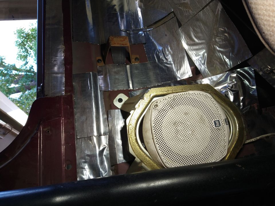Vehicle interior with a white speaker and gold trim, covered in silver sound deadening material.