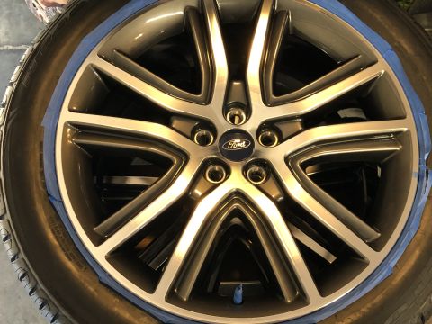 2019 Ford Everest - A close-up view of a gray and silver 5-spoke alloy wheel with a blue Ford logo in the center. The wheel is taped off with blue painter's ta…
