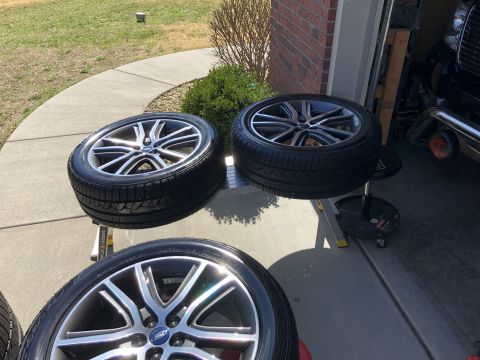 2019 Ford Everest - Four aftermarket alloy wheels with tires are displayed on a metal stand and a rolling stool in front of a garage. The wheels have a multi-s…