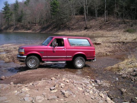 1994 Ford Bronco - A red Ford Bronco navigates a rocky stream bed in a wooded area.