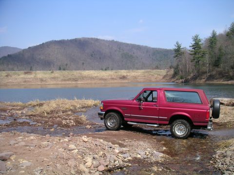 1994 Ford Bronco - A red 1994 Ford Bronco is parked near the water's edge with a wooded hill in the background.