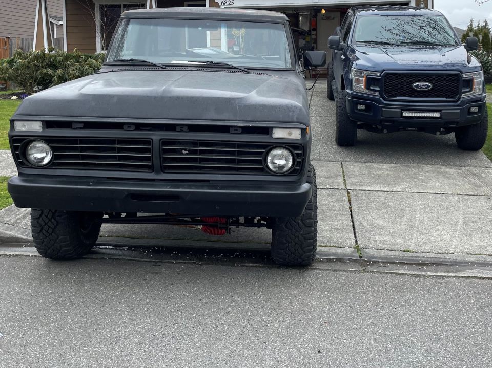 Black 1978 Ford Bronco front view, lifted with large tires, parked in driveway next to a blue Ford F-150
