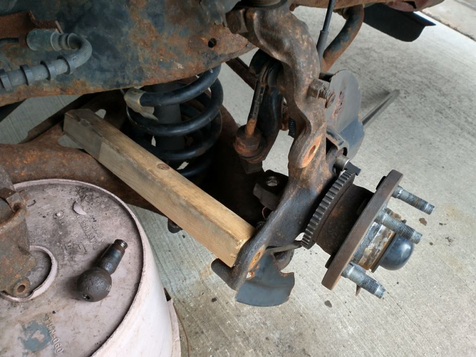 To free a stuck ball joint stud, a pipe wrench (not pictured) is clamped onto and used to turn the ball via the added cheater bar.  The steering knuckle is braced in place by a piece of wood inserted between the trailing edge of the knuckle and the cut-out in the lower control arm.