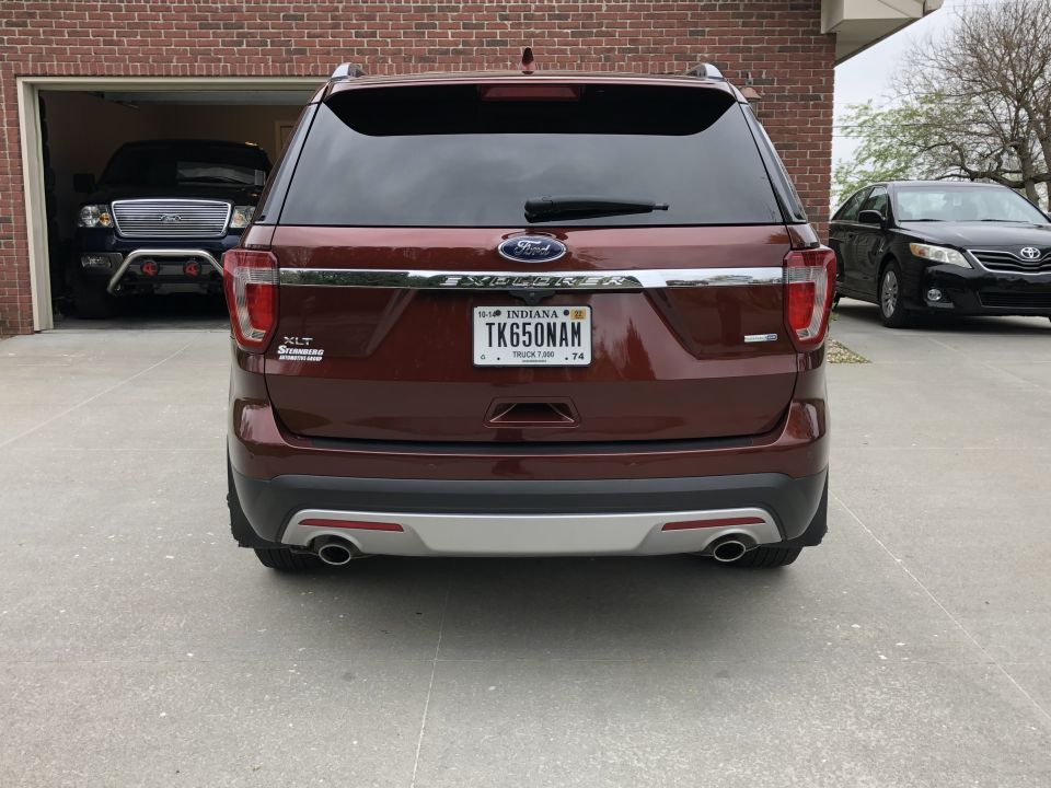 Rear view of a maroon Ford Explorer with Indiana plates. A black sedan is parked to the right, and a dark pickup truck is in the garage behind it