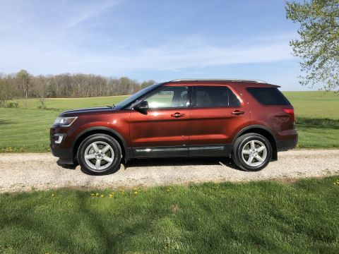2016 Ford Explorer - This orange SUV is parked on a gravel road with a grassy field and trees in the background under a clear blue sky.