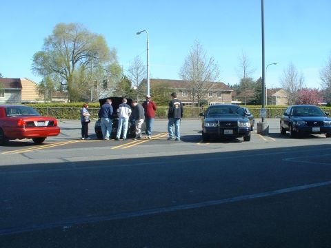 1999 Ford Crown Victoria - Several people are gathered around the open hoods of cars in a parking lot on a sunny day.