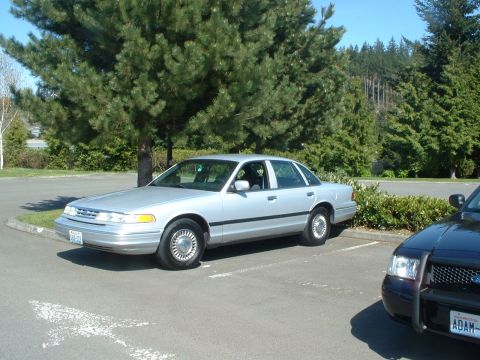 1999 Ford Crown Victoria - A silver 1999 Ford Crown Victoria is seen parked in a lot, with a police vehicle partially visible in the foreground.