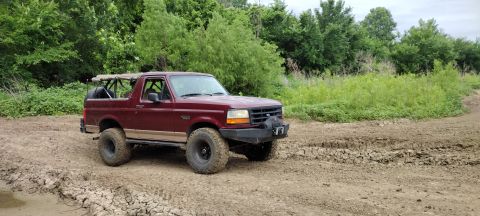 1996 Ford Bronco - A maroon 1996 Ford Bronco is parked on a muddy track surrounded by greenery.