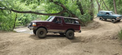 1996 Ford Bronco - Two Ford Broncos navigate a wooded off-road trail with sandy terrain.