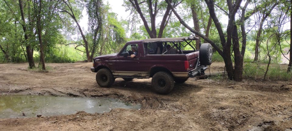 Maroon 1996 Ford Bronco with soft top removed, driving on a dirt trail with mud puddles