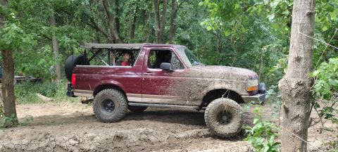 1996 Ford Bronco - maroon and tan Ford Bronco with its top removed and muddy off-road tires climbs a dirt trail through wooded terrain.