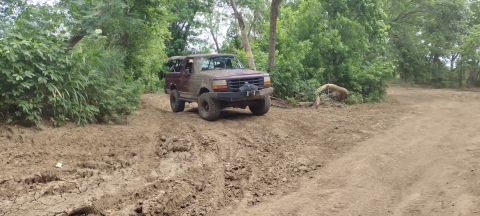 1996 Ford Bronco - A maroon Ford Bronco navigates a muddy off-road trail surrounded by trees.