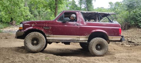 1996 Ford Bronco - A red 1996 Ford Bronco with off-road tires navigates a muddy trail in a wooded area.