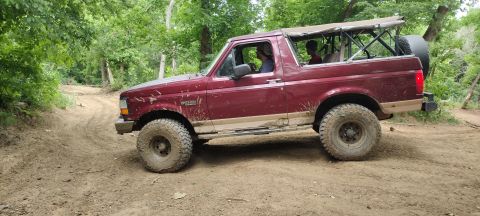 1996 Ford Bronco - A maroon 1996 Ford Bronco navigates a dirt path surrounded by trees, its soft top revealing passengers.