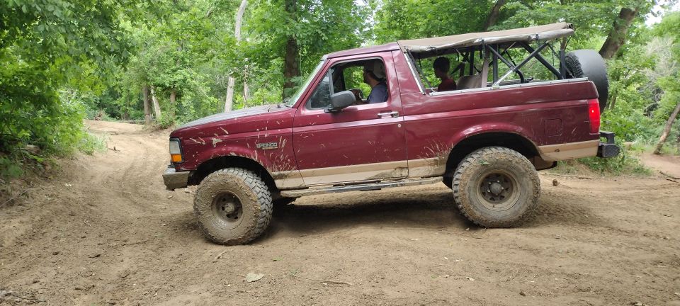Maroon 1996 Ford Bronco with a soft top driving on a dirt trail, showing mud splatter and off-road tires