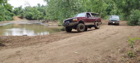 1996 Ford Bronco - Two Ford Broncos navigate a muddy off-road trail with trees and water nearby.