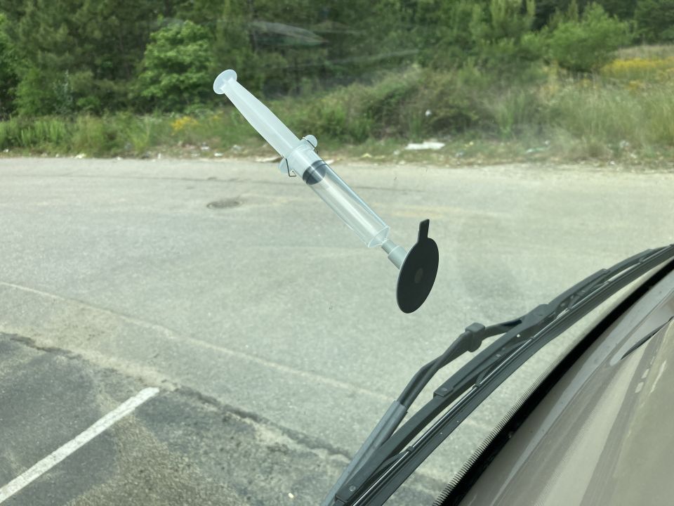 Clear plastic syringe suction-cup tool stuck to a windshield with parking lot in background