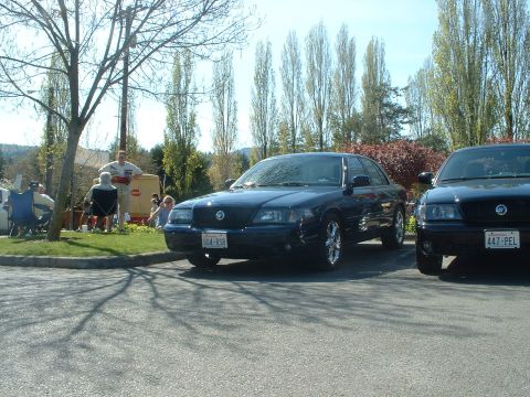 1999 Ford Crown Victoria - Two dark blue sedans are parked side-by-side in a lot with people gathered in the background.