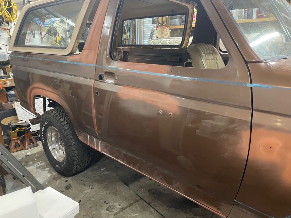 Side view of a brown 1984 Ford Bronco with primer showing, in a garage