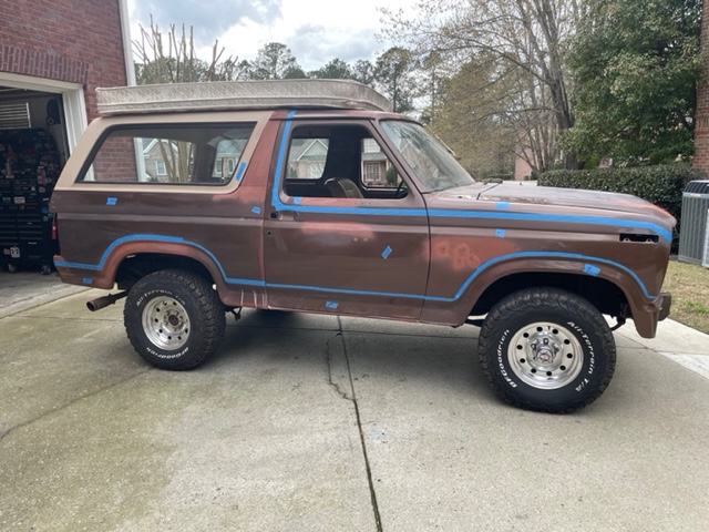 Brown Ford Bronco with blue trim, showing primer spots and a mattress on the roof