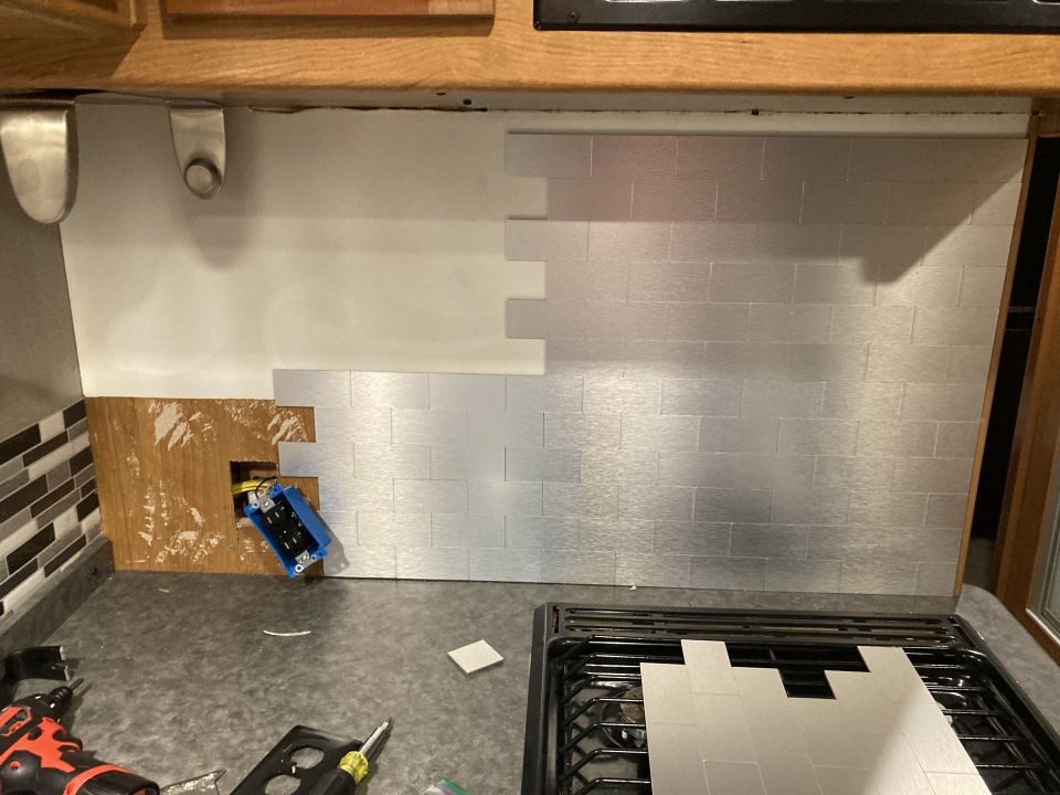 Backsplash with silver subway tiles being installed over wood paneling, exposing an electrical box