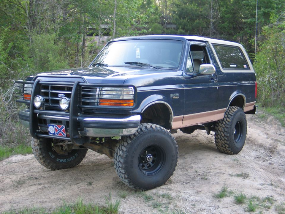 Lifted dark blue and tan Ford Bronco with brush guard and mud tires on a sandy wooded trail