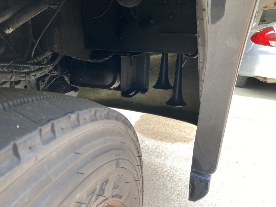 Air horns mounted under a vehicle chassis, near a large tire. Another vehicle is partially visible to the right