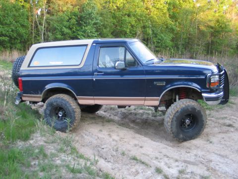 1995 Ford Bronco - A lifted blue Ford Bronco with off-road tires is parked on a dirt path surrounded by trees.