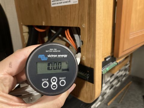 2015 Ford F-550 - A hand holds a Victron BMV-702 battery monitor displaying 100.0% charge, with wiring and cabinetry in the background.