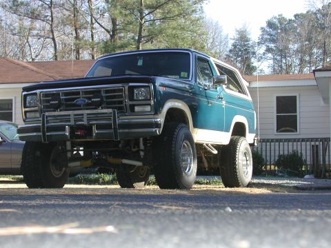 1985 Ford Bronco - Lifted 1985 Ford Bronco in teal and white, seen from a low front 3/4 angle.