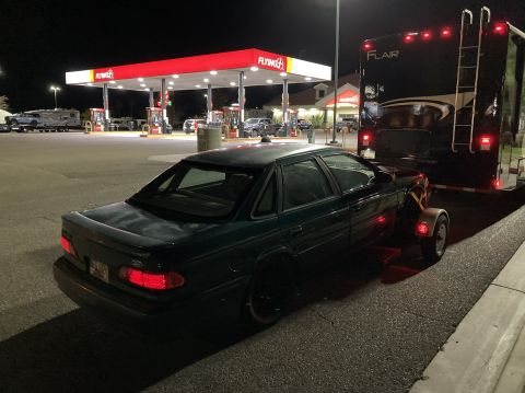 2015 Ford F-550 - A dark sedan is seen being towed by a dolly at a gas station after dark, with a large RV trailer behind it.