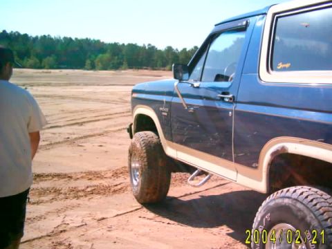 1985 Ford Bronco - A blue and white 1985 Ford Bronco is parked on a sandy surface, possibly at a clay pit, with a person standing nearby.