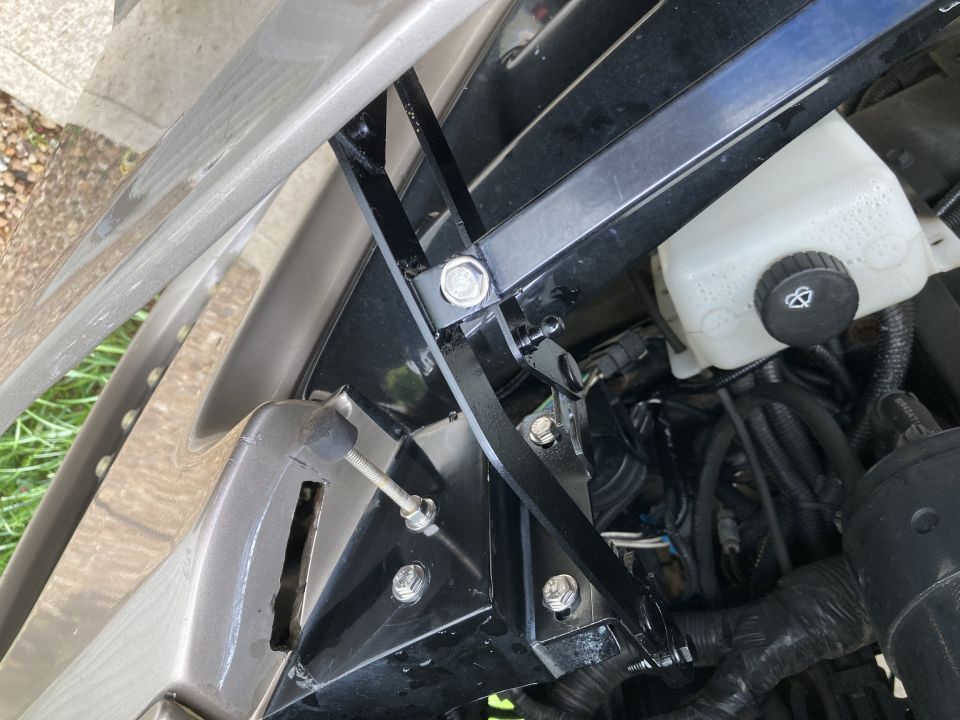 Close-up of a black metal hood bracket assembly with bolts and a white fluid reservoir