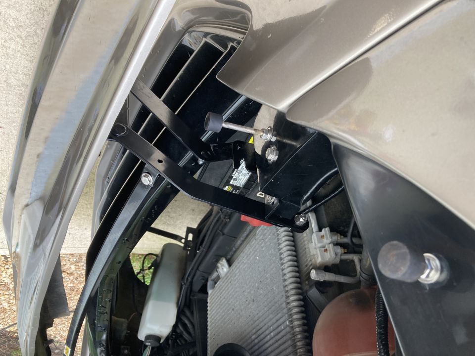 Close-up of a black metal hood bracket assembly attached to a vehicle's grille and fender