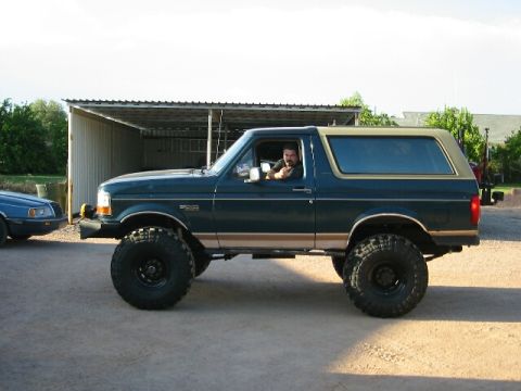 1994 Ford Bronco - A dark green Ford Bronco with a tan top and lifted suspension is parked outside a garage.